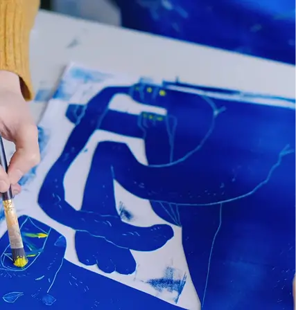 A person in a yellow sweater, inspired by Flying Arts Alliance, uses a paintbrush to add yellow details to a blue artwork featuring abstract shapes and figures on a table.