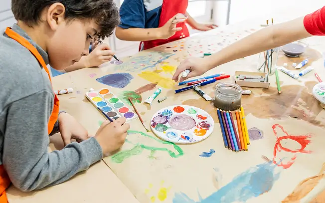 A group of primary school students paint together at a table while an artist's hand reaches in to help