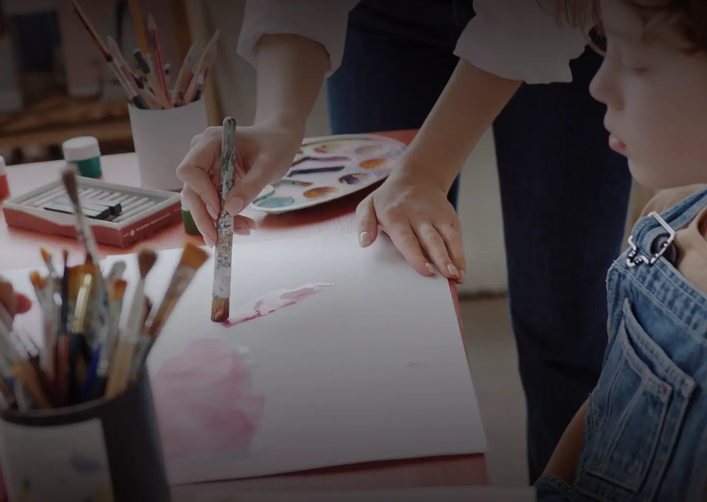 An adult demonstrates watercolor painting on paper while a child in denim overalls watches closely. Paintbrushes, a palette, and Flying Arts Alliance art supplies are spread across the table.