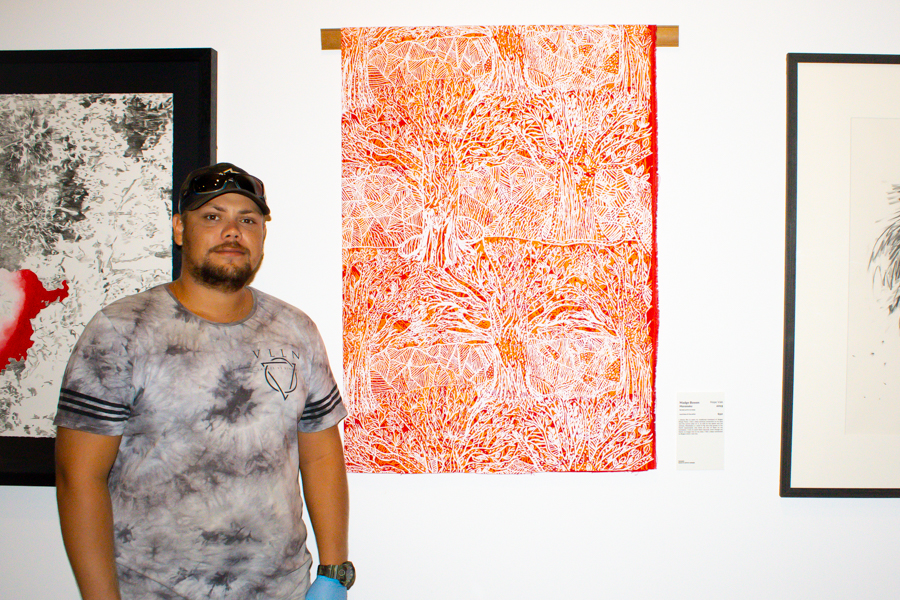 Jaivan is smiling next to an artwork made by his grandmother Madge Bowen. The artwork is an intricate red, orange and white screen print.